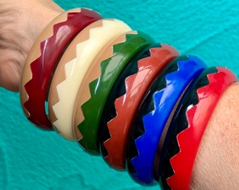 Può includere: Una collezione di braccialetti colorati in varie tonalità, tra cui rosso, beige, verde, marrone, blu e rosso. Ogni braccialetto presenta un motivo a zig-zag in un colore a contrasto. I braccialetti sono impilati su un polso.