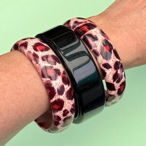 Op de afbeelding: Een set van drie armbanden bestaande uit twee bijpassende armbanden met een rood en zwart luipaardpatroon en één effen glanzend zwarte armband in het midden. De armbanden zijn glad, afgerond en hebben een gepolijste afwerking.