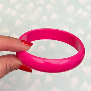 Pu&ograve; includere: Un braccialetto bangle rosa acceso. La superficie liscia e lucida riflette la luce, evidenziando il suo colore pieno. Questo accessorio rotondo e audace &egrave; un pezzo forte, perfetto per aggiungere un tocco di colore a qualsiasi outfit.