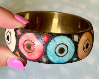 Può includere: Un braccialetto a cerchio in ottone con una fascia nera con disegni circolari colorati in bianco, rosso e blu. I cerchi hanno un punto centrale dorato e sono accentuati da piccole forme quadrate e rettangolari.
