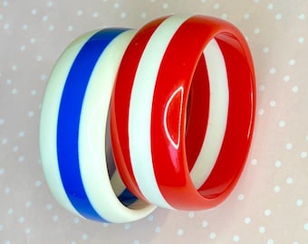 Può includere: Due braccialetti colorati. Uno è bianco con strisce blu, l'altro è rosso con strisce bianche. I braccialetti sono realizzati in materiale lucido e sono impilati. Lo sfondo è rosa chiaro con pois bianchi.
