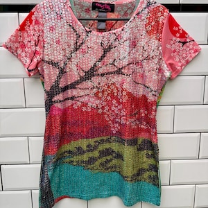 Op de afbeelding: Een T-shirt met korte mouwen en een levendige landschapsprint met kersenbloesembomen, een rode zon en groene heuvels. Het gehele vooroppervlak is bedekt met kleine, glinsterende pailletten. Het label in de nek bevat de tekst Custo Barcelona.