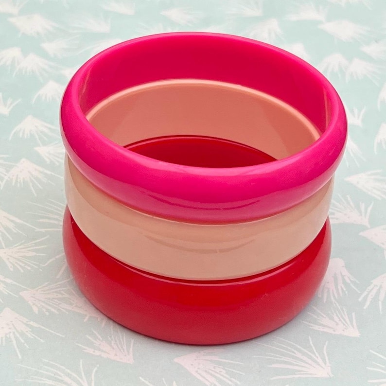 Pu&ograve; includere: Tre braccialetti impilati nelle tonalit&agrave; del rosa e del rosso. Il braccialetto superiore &egrave; un fucsia vibrante, quello centrale &egrave; un pesca tenue e quello inferiore &egrave; rosso intenso. I braccialetti sono lisci e lucidi, creando un accessorio audace e colorato.