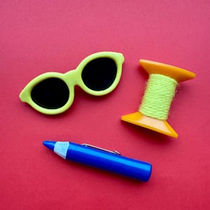 Op de afbeelding: Een stilleven op een rode achtergrond met een limoengroene zonnebril, een blauwe pen met een zilveren clip en een gele spoel met groen garen. De zonnebril heeft zwarte glazen.