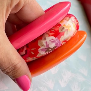 Könnte beinhalten: Drei farbenfrohe Armreifen. Einer ist ein leuchtendes Pink, ein anderer ein leuchtendes Orange, und der dritte hat ein Blumenmuster mit roten, rosa und gelben Blumen. Die Armreifen sind glatt und glänzend.