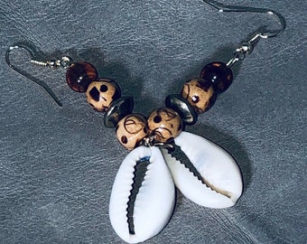 May include: Handmade earrings featuring a combination of natural elements. The earrings have silver-tone hooks, wooden beads with carved designs, and polished white cowrie shells. The beads are brown and tan, and the shells are white.