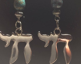 May include: A pair of silver-toned earrings featuring a Chai symbol. Each earring has a turquoise and black teardrop-shaped bead, and a silver-toned bead above the Chai symbol. The earrings hang from a silver-toned hook.