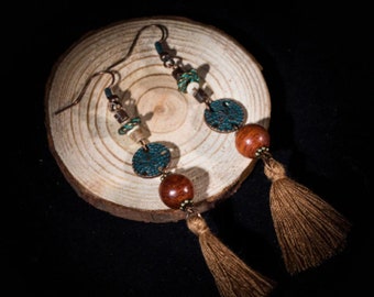 May include: A pair of dangle earrings with a bohemian design. Each earring features a copper-colored disc, a wooden bead, and a brown tassel. The earrings are set against a wooden slice on a black background.