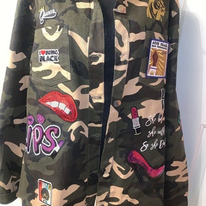 May include: A camouflage jacket with various patches, including a red lip, a crown, a black heart with the words "I BEING BLACK", a purple "lips" patch, a black and white afro with the word "SWAG", a lion, a raised fist with the words "ARISE ISRAEL QAN YAGHARARIA 144", a red high heel, and a lipstick with the text "She believe she walk & she Do".