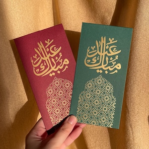 May include: Two greeting cards, one burgundy and one green, with gold calligraphy and a decorative floral pattern. The cards are rectangular and feature Arabic script. The background is a golden-colored fabric.