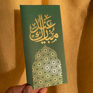 May include: A dark green card with gold Arabic calligraphy and a decorative gold geometric pattern. The card is held against a gold fabric background. The calligraphy reads "Eid Mubarak".
