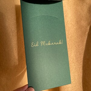 May include: A dark green envelope with the gold text "Eid Mubarak!". The envelope is rectangular and has a curved flap. The background is a light gold color.