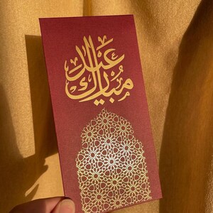 May include: A burgundy card with gold Arabic calligraphy and a decorative gold floral pattern. The card is rectangular and held by a hand, with the text "Eid Mubarak" in gold script.