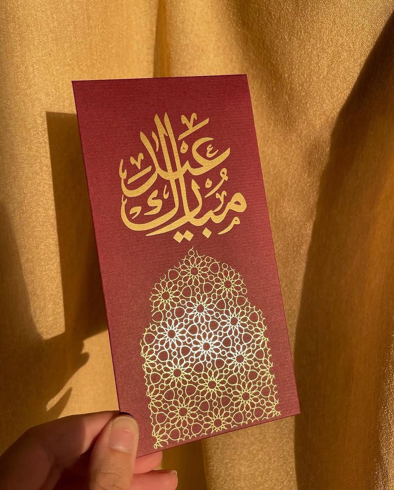May include: A burgundy card with gold Arabic calligraphy and a decorative gold floral pattern. The card is rectangular and held by a hand, with the text "Eid Mubarak" in gold script.