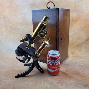 May include: An antique microscope with brass components and a black base, next to a wooden box and a Coca-Cola can. The microscope has multiple lenses and adjustment knobs. The Coca-Cola can is red with white lettering.