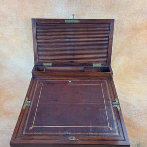 Peut inclure: Un bureau d'écriture antique en bois avec un dessus en cuir et des ferrures en laiton. Le bureau est ouvert et l'intérieur est visible, montrant une série de compartiments et un couvercle coulissant.