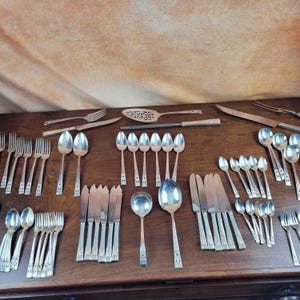 Puede incluir: Un juego de cubiertos vintage, que incluye tenedores, cucharas y cuchillos, dispuestos sobre una superficie de madera. Los cubiertos están hechos de plata pulida y tienen un diseño clásico.