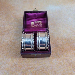 May include: Two silver napkin rings with a beaded edge are displayed in a vintage purple velvet-lined box. The box is open, revealing the rings and the maker's mark. The rings are cylindrical and have a shiny finish.