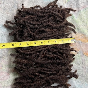 May include: A bundle of dark brown dreadlocks, approximately 9 inches long, is displayed against a textured background. The dreadlocks are tightly coiled and have a natural, textured appearance. A yellow measuring tape is placed across the dreadlocks.