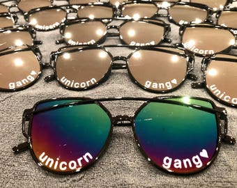 May include: A collection of black-framed sunglasses with iridescent lenses. Each pair features white text printed on the lenses that reads "unicorn" on the left and "gang" with a small heart symbol on the right. The frames have a double bridge.