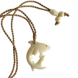 May include: Cream-colored shark pendant necklace with a braided tan cord. The shark pendant is carved with details like gills and a small eye. The necklace has a sliding bead closure for adjustable length.