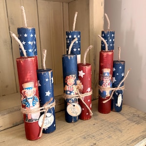 May include: Hand-painted wooden firecracker decorations in red and blue with white stars. Each firecracker has a jute wick and is adorned with patriotic images and a tag that reads "4th July". These decorative pieces are perfect for Independence Day.