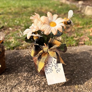 May include: A small, rustic metal pot filled with artificial daisies and greenery, adorned with a plaid bow. Miniature bee figurines are attached to the flowers. A tag reads "Save The Bees".