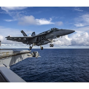 May include: A grey fighter jet with black markings is landing on an aircraft carrier. The jet is in the air, with the landing gear down. The carrier is in the ocean, with blue water and white clouds in the background.
