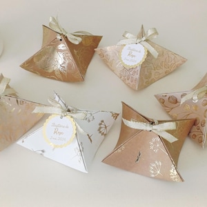 Peut inclure: Six boîtes cadeaux décoratives en forme de pyramide, dans des tons or, rose et blanc, ornées de motifs floraux et botaniques. Chaque boîte est nouée avec un ruban crème. Certaines boîtes ont une petite étiquette avec du texte.