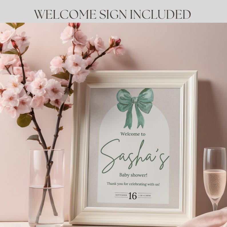 Pu&ograve; includere: Un cartello di benvenuto per un baby shower incorniciato con un'illustrazione di fiocco verde. Il cartello recita "Welcome to Sasha's Baby shower!" con la data "SEPTEMBER 16". Un vaso di fiori rosa e un calice di champagne sono sullo sfondo.