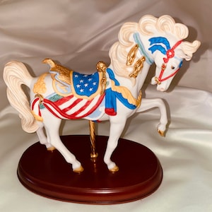 May include: A white porcelain carousel horse figurine with a red, white, and blue American flag saddle and blue and gold ribbons. The horse is standing on a wooden base.