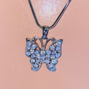 Puede incluir: Un collar de cadena de plata con un colgante de mariposa. La mariposa está cubierta de diamantes de imitación transparentes.