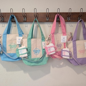 May include: Four canvas tote bags in blue, green, pink, and purple, each with a matching mini tote and a Trader Joe's logo. The bags hang from a wooden rack against a white wall. Each bag has a cream-coloured centre panel.