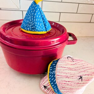 Może przedstawiać: Magenta żeliwny garnek z pasującą pokrywką, zwieńczony niebiesko-białym wzorzystym uchwytem do garnka. W pobliżu znajduje się kolejna podkładka pod garnek z różowo-białym wzorem i żółtym wykończeniem. Uchwyty do garnków mają kształt stożka.