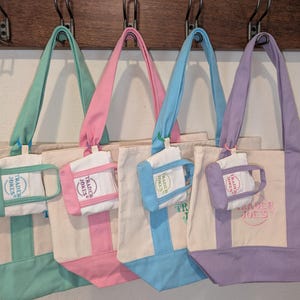 May include: Four canvas tote bags in pastel colors: mint green, pink, light blue, and lavender. Each bag has a matching colored handle and base, with a small, attached pouch. The bags are hanging on a wooden rack.