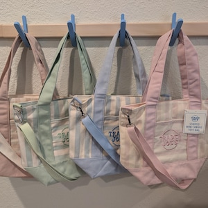 May include: Four striped mini canvas tote bags with shoulder straps, each featuring a Trader Joe's logo. The bags come in pink, green, blue, and a lighter pink, all with matching solid-colored bases and handles. A product tag is attached to the pink bag.