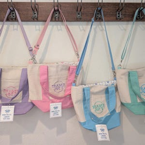 Może przedstawiać: Cztery mini płócienne torby w pastelowych kolorach lawendy, różu, błękitu i turkusu, każda z pasującym paskiem i logo Trader Joe's. Torby wiszą na drewnianym wieszaku. Do pierwszej torby przymocowana jest mała torba Trader Joe's.