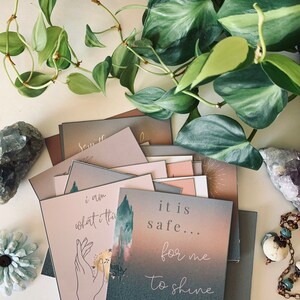 May include: A stack of cards with inspirational quotes. The top card is a light blue color with the text "it is safe... for me to shine". The cards are on a white surface with a green plant and crystals.