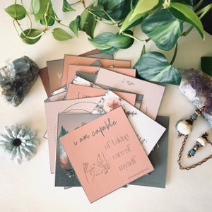 May include: A stack of pink and gray affirmation cards with the text "I am capable of taking care of myself" on the top card. The cards are decorated with floral patterns and a hand drawn illustration of hands holding a circle.