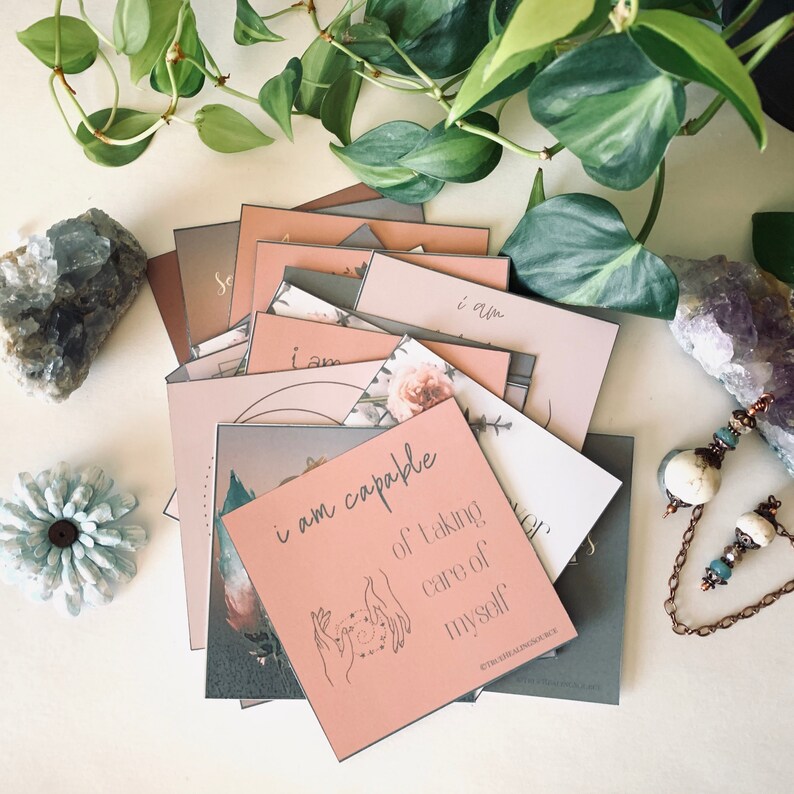 May include: A stack of pink and gray affirmation cards with the text "I am capable of taking care of myself" on the top card. The cards are decorated with floral patterns and a hand drawn illustration of hands holding a circle.