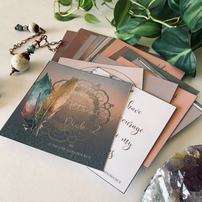 May include: A stack of Law of Attraction affirmation cards with a gray card on top featuring a mandala design and two feathers. The text on the gray card reads "Law of Attraction Deck 2 by @TRUEHEALINGSOURCE".