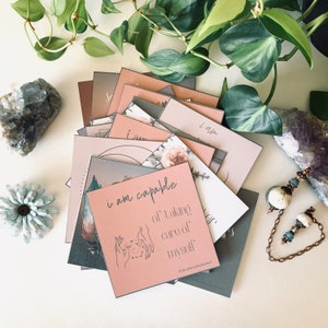 May include: A stack of pink and gray affirmation cards with the text "i am capable of taking care of myself" on the top card. The cards are decorated with floral patterns and a hand drawing of a person holding a circle with stars.
