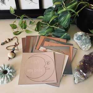 May include: A stack of affirmation cards with a pink card on top featuring a crescent moon with a face and the text "i love my body". The card is surrounded by other cards with various colors and designs. The cards are on a white surface with a green plant, a blue crystal, and a purple crystal.