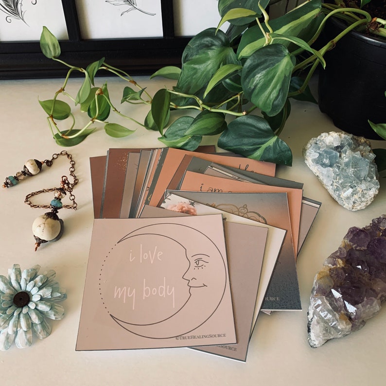 May include: A stack of affirmation cards with a pink card on top featuring a crescent moon with a face and the text "i love my body". The card is surrounded by other cards with various colors and designs. The cards are on a white surface with a green plant, a blue crystal, and a purple crystal.
