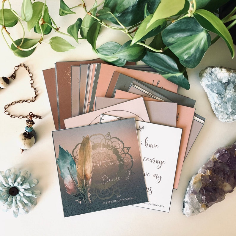May include: A set of Law of Attraction affirmation cards with a pink, brown, and gray color scheme. The cards feature a mandala design with feathers and the text "Law of Attraction Deck 2" on the front. The cards are arranged in a pile on a white surface with crystals and a pendulum.