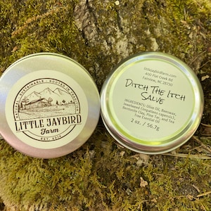 May include: A silver tin with a green and white label that reads "Little Jaybird Farm Est. 2014" and a silver tin with a green and white label that reads "Ditch The Itch Salve" with the ingredients listed. The tins are sitting on a bed of green moss.