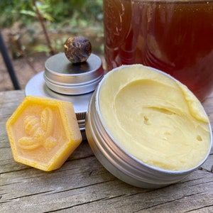 May include: A close-up of a honey-themed gift set. The set includes a small jar of honey, a round container of beeswax balm, and a hexagonal beeswax candle with a bee design.