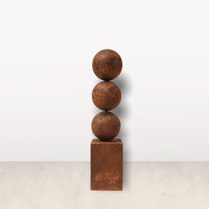 Può includere: Una scultura rustica con tre sfere impilate su una base quadrata. La scultura ha un aspetto strutturato e invecchiato con un colore marrone-rossastro, che suggerisce un'estetica esterna o industriale. Il design è minimalista e moderno.