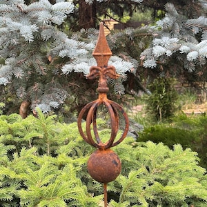 May include: A weathered metal garden finial with a pointed top, decorative flourishes, and a spherical base. The finial has a cage-like design and is set against a backdrop of green and blue-green foliage.