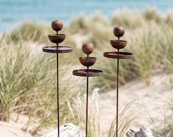 May include: Three tall, thin metal garden stakes with a rusted finish. Each stake features a vertical arrangement of a flat circular tray, a bowl-shaped component, and a spherical topper, all supported by a slender rod anchored in the ground.